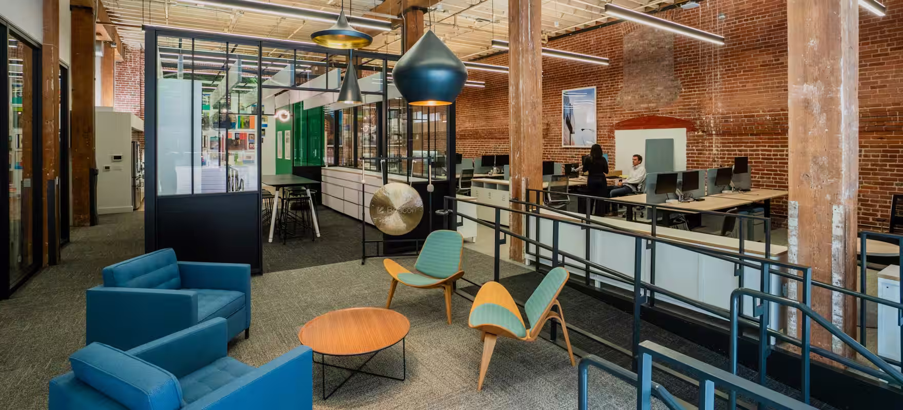 Inside Beacon&rsquo;s office: a converted brick loft with timber columns and pendant lamps, a lounge in front, and rows of desks beyond.